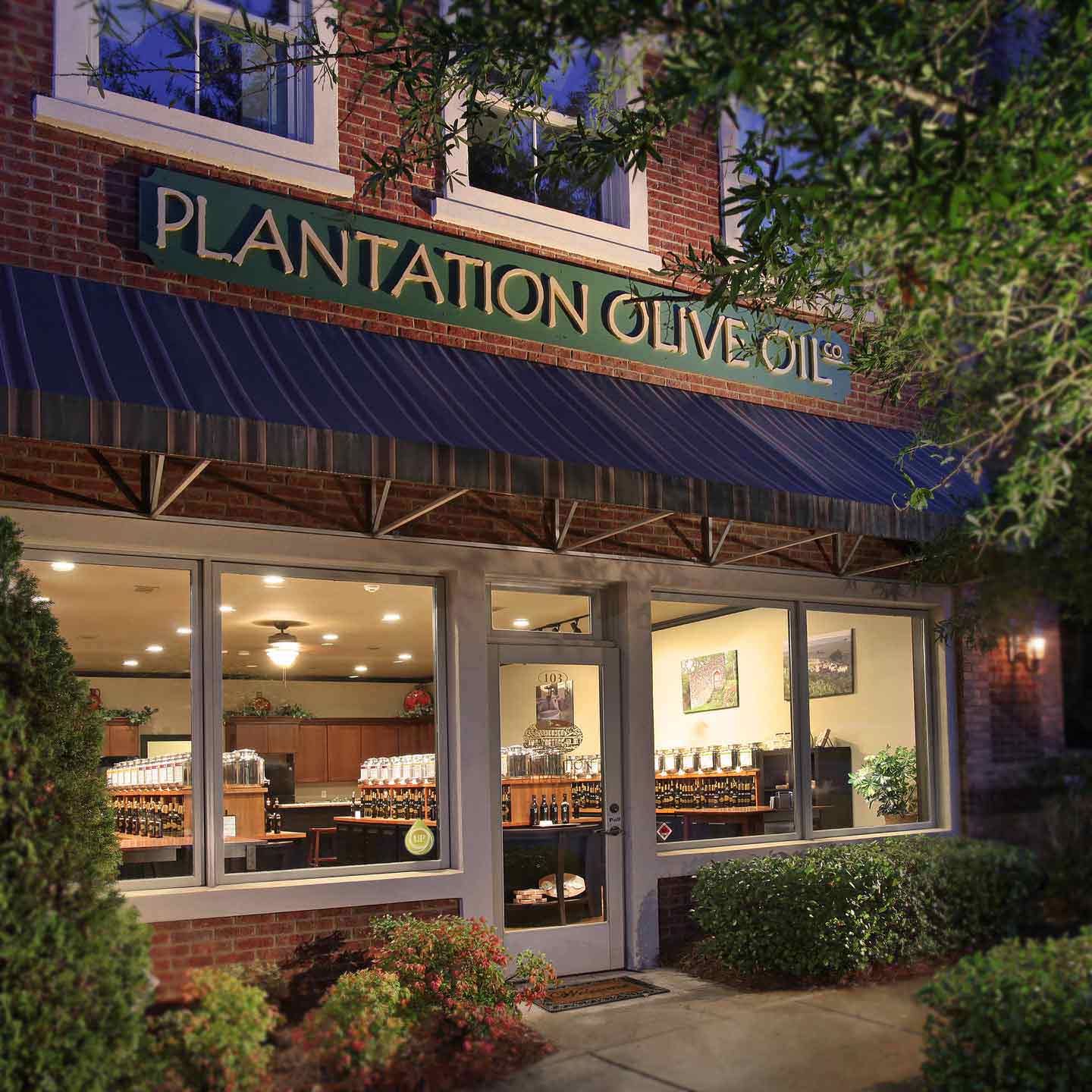 Nighttime exterior photograph showing the illuminated interior of the olive oil store.