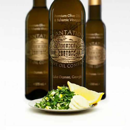 Photo showing fresh minced garlic, mint and parsley on a dish with 2 lemon slices in front of three bottles of olive oil.