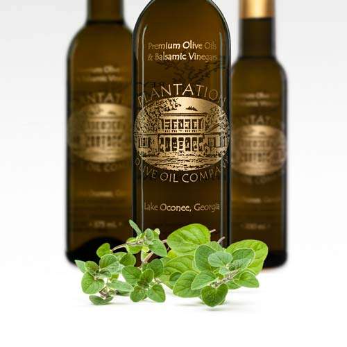 Sprigs of fresh oregano lay in front of three gourmet olive oil bottles on a white background.