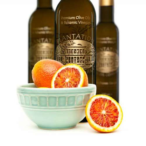 Photo showing a porcelain bowl of fresh whole and cut blood oranges in front of three bottles of olive oil.