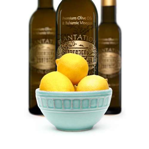 Photo showing light blue porcelain bowl filled with whole eureka lemons in front of three bottles of olive oil